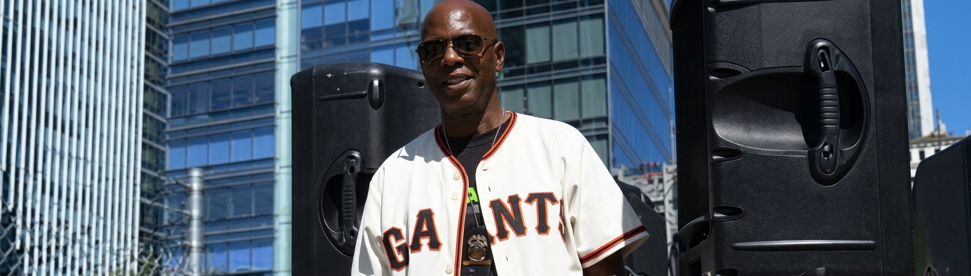 John Parker in a San Francisco Giants jersey and sunglasses, standing between PA speakers with city buildings behind him